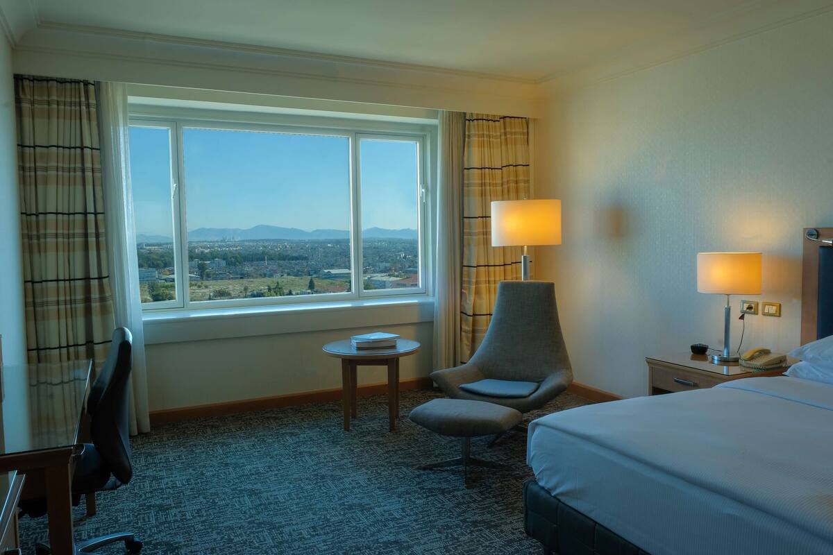 A hotel room with a large bed, two bedside lamps, a chair, a small table, and a view of a cityscape through a large window.
