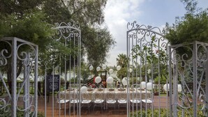 Un área de comedor al aire libre con mesas y sillas blancas, rodeada por puertas de metal ornamentadas y follaje.