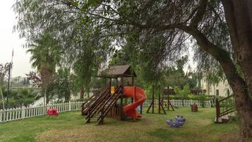 Children can play on the playground's wooden structure, swings, and climbing frame, surrounded by lush greenery and a white picket fence.