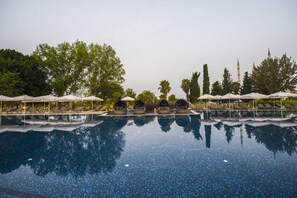 A swimming pool with lounge chairs and umbrellas, surrounded by trees and a mosque in the background. - Adana Hilton SA (Adana)