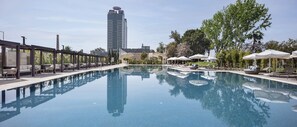 A swimming pool with lounge chairs and umbrellas, surrounded by trees and a tall building in the background. - Adana Hilton SA (Adana)