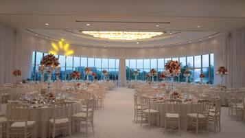 A large, elegantly decorated banquet hall with round tables and chairs, large windows, and a chandelier.