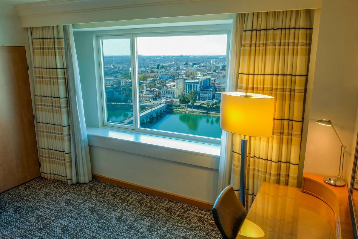 A hotel room with a large window offering a city view, a desk with a lamp, and a chair.
