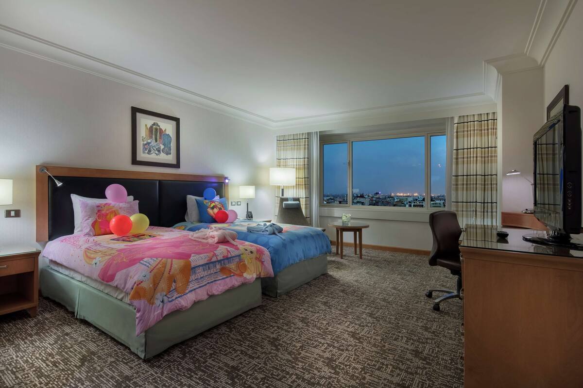 A hotel room with two beds, a desk, a chair, and a large window offering a city view.