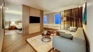 Una habitación de hotel moderna con sofá, una mesa de centro, un televisor de pantalla plana y vistas a la ciudad a través de amplios ventanales.