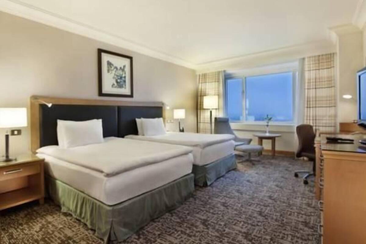 A hotel room with two beds, a desk, a chair, a window with a view, and a framed picture on the wall.