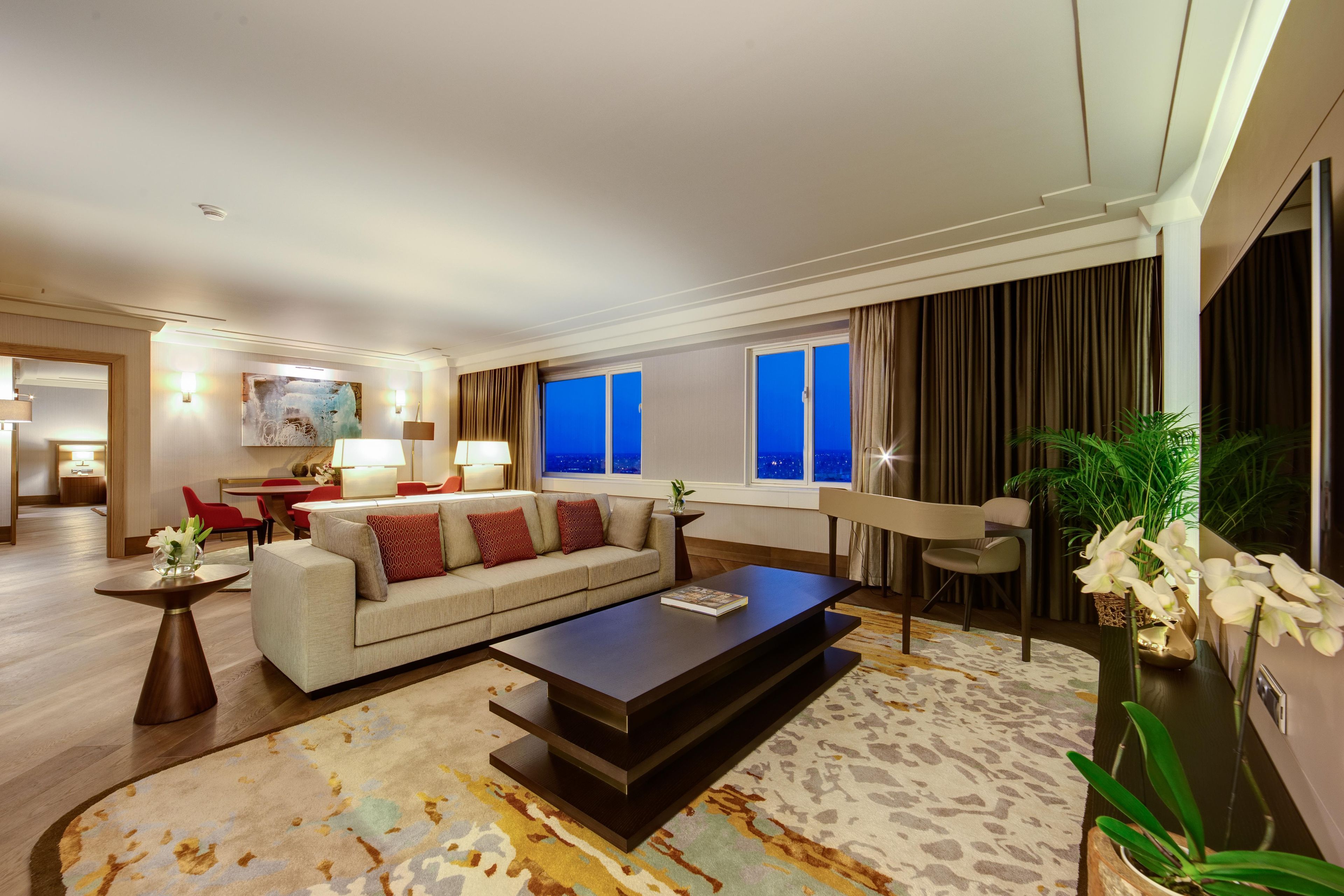 A modern hotel room with a sofa, a coffee table, a dining area, and a view of the city at night.
