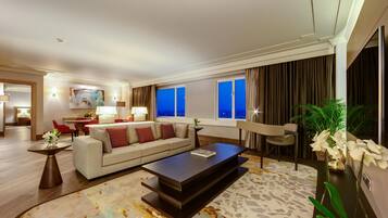 A modern hotel room with a sofa, a coffee table, a dining area, and a view of the city at night.