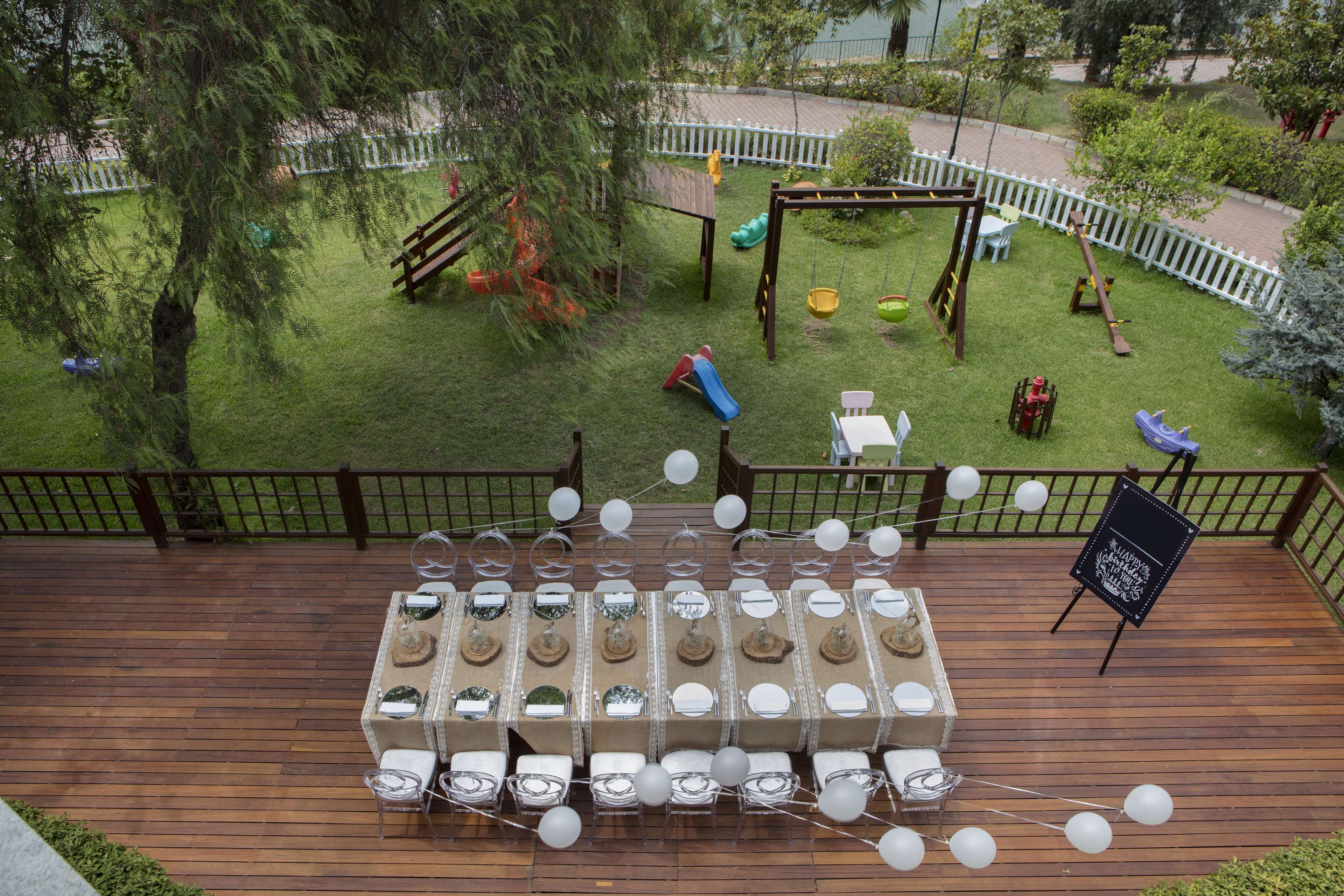 Une terrasse avec des tables préparées pour une fête, entourée d’une autre terrasse en bois et d’un jardin clos avec des jeux pour enfants.