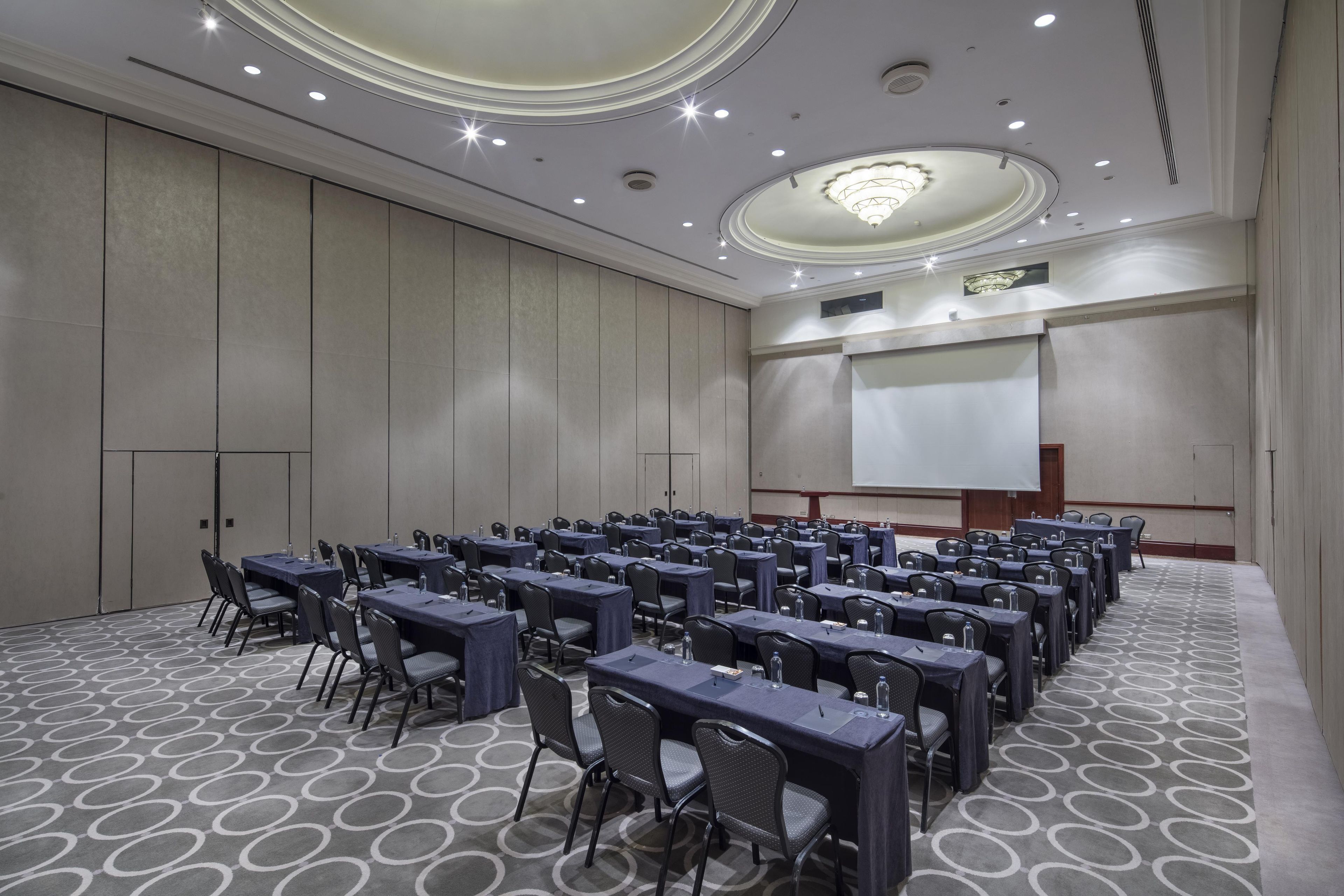 Une salle de conférence avec des rangées de tables et de chaises, un écran de projection et une scène.