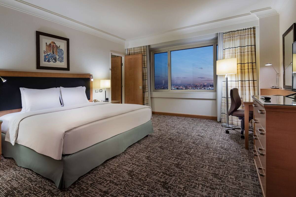 A hotel room with a large bed, a desk with a chair, a television, and a window with a city view.