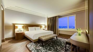 A hotel room with a large bed, bedside lamps, a bench, a small table with a vase, and a view of the city through the window.
