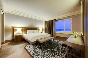 Habitación de hotel con una cama grande, lámparas de noche, un banco, una mesita con un florero y vistas a la ciudad a través de la ventana.