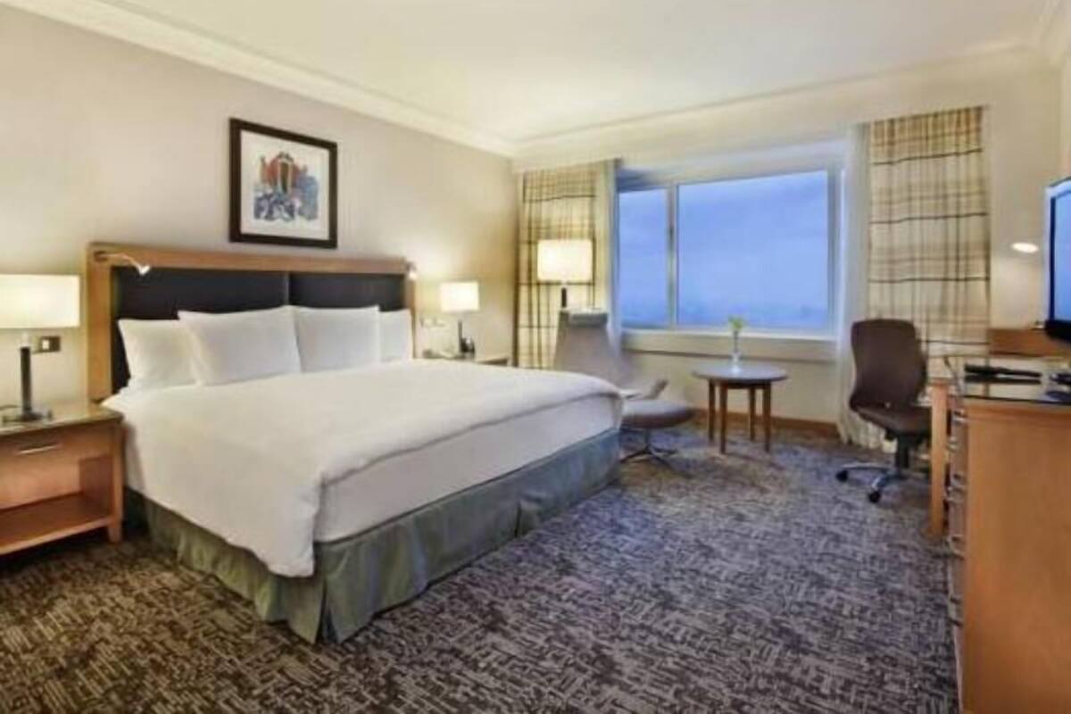 A hotel room with a large bed, bedside tables, a desk, a chair, a window with curtains, and a painting on the wall.