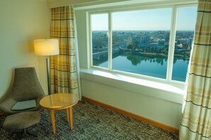 A hotel room with a large window offering a view of a cityscape and a river. There is a round table and two armchairs. - Adana Hilton SA (Adana)
