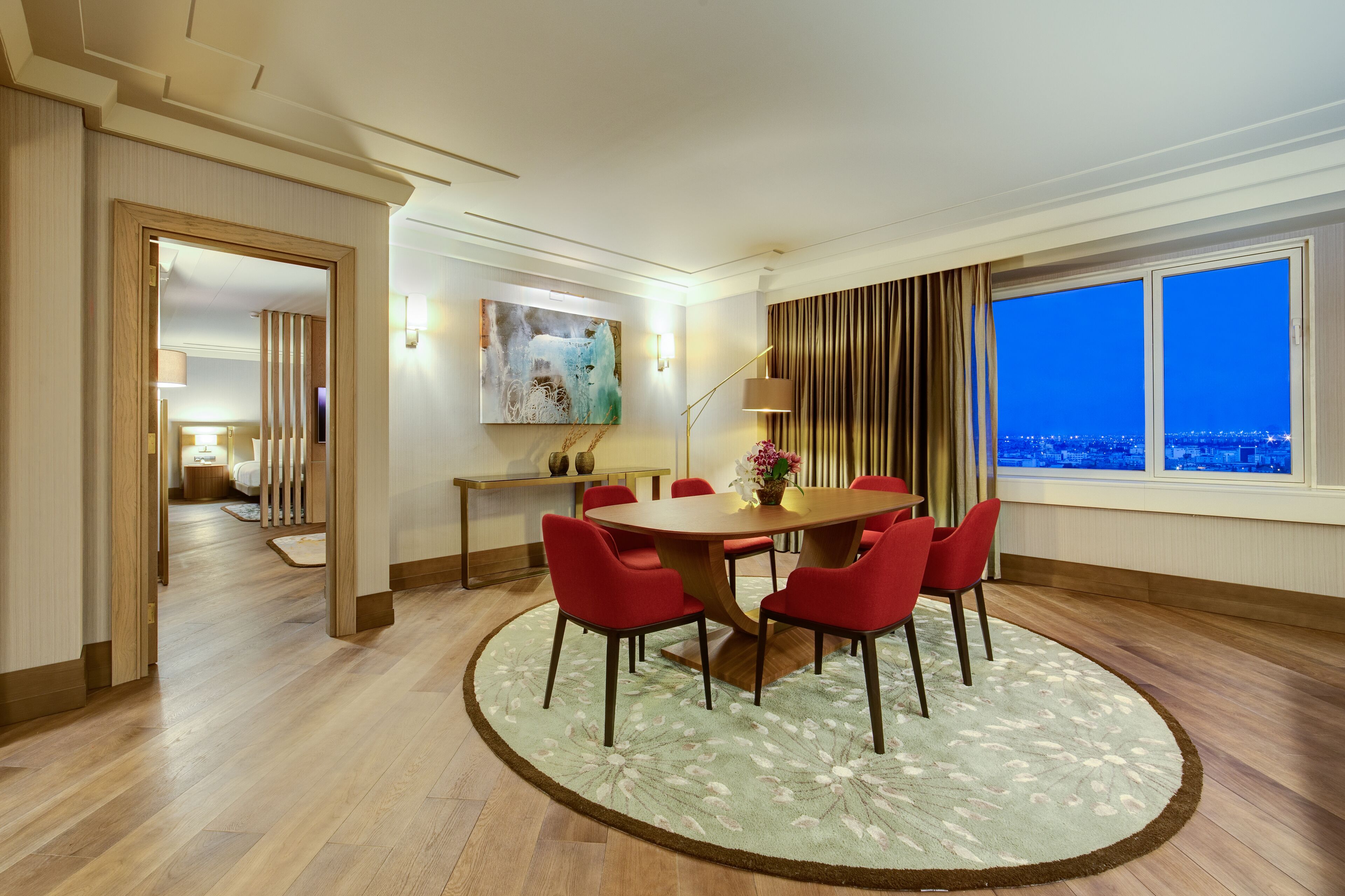 A hotel room with a dining area featuring a wooden table and red chairs, a large window with a city view, and a modern art piece on the wall.