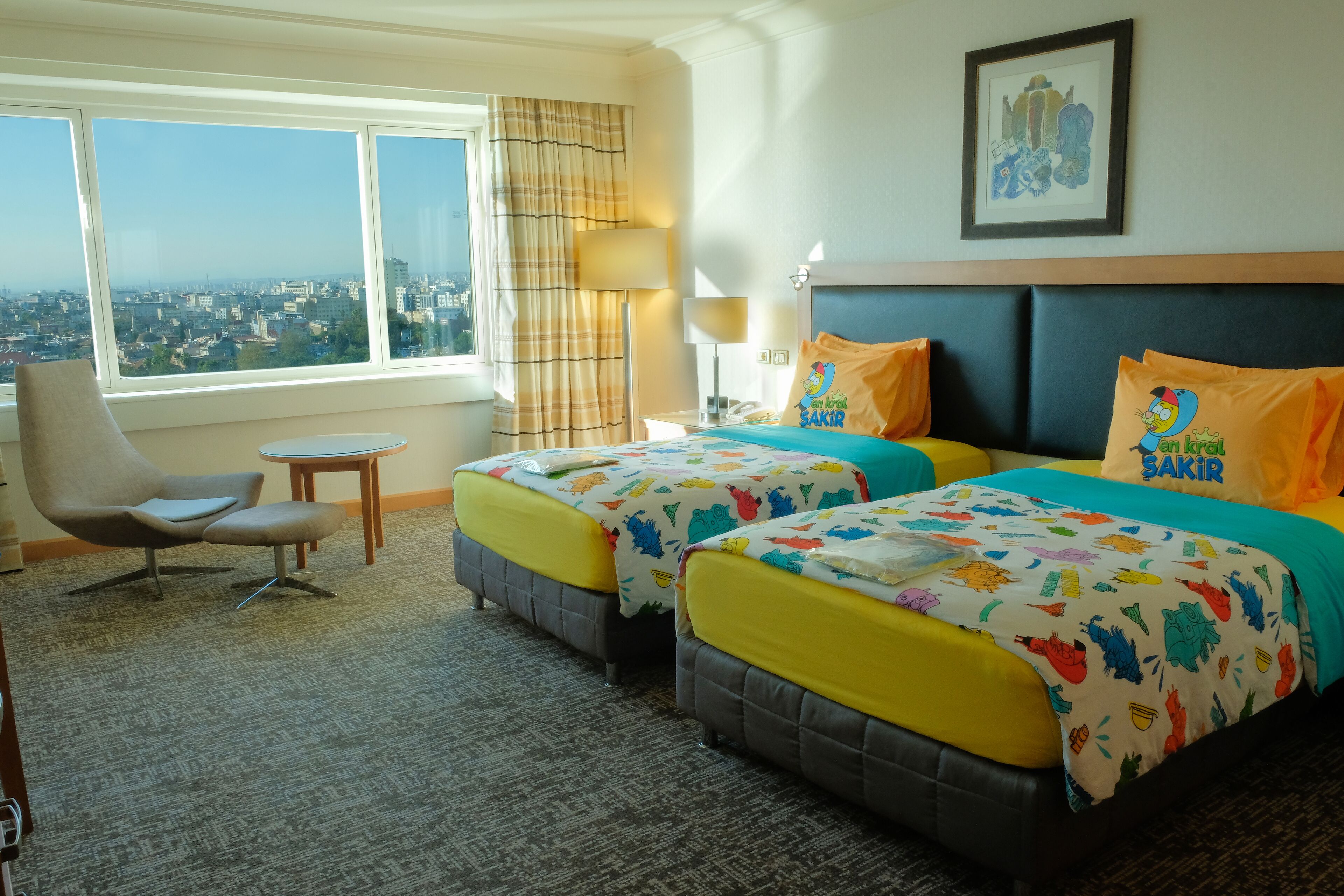 Een hotelkamer met twee bedden, een stoel, een tafeltje en uitzicht op de stad.