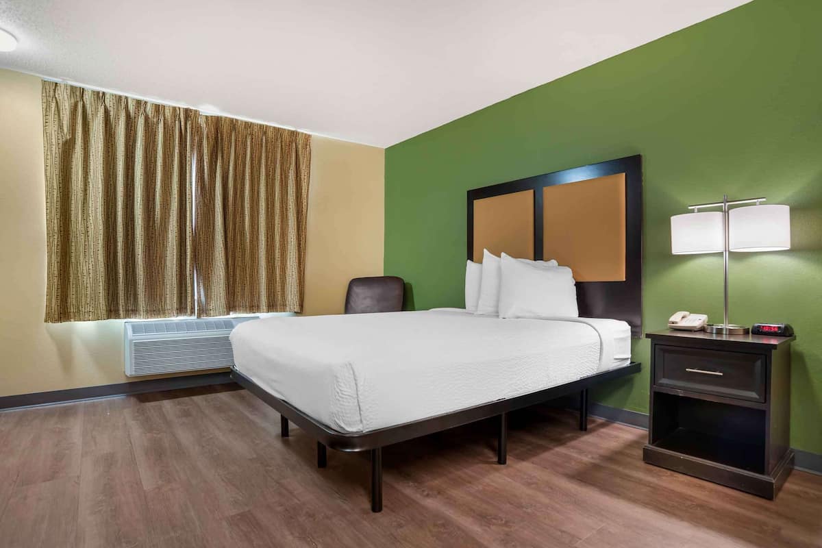 A hotel room with a bed, a chair, a nightstand with a lamp, and a window with curtains.