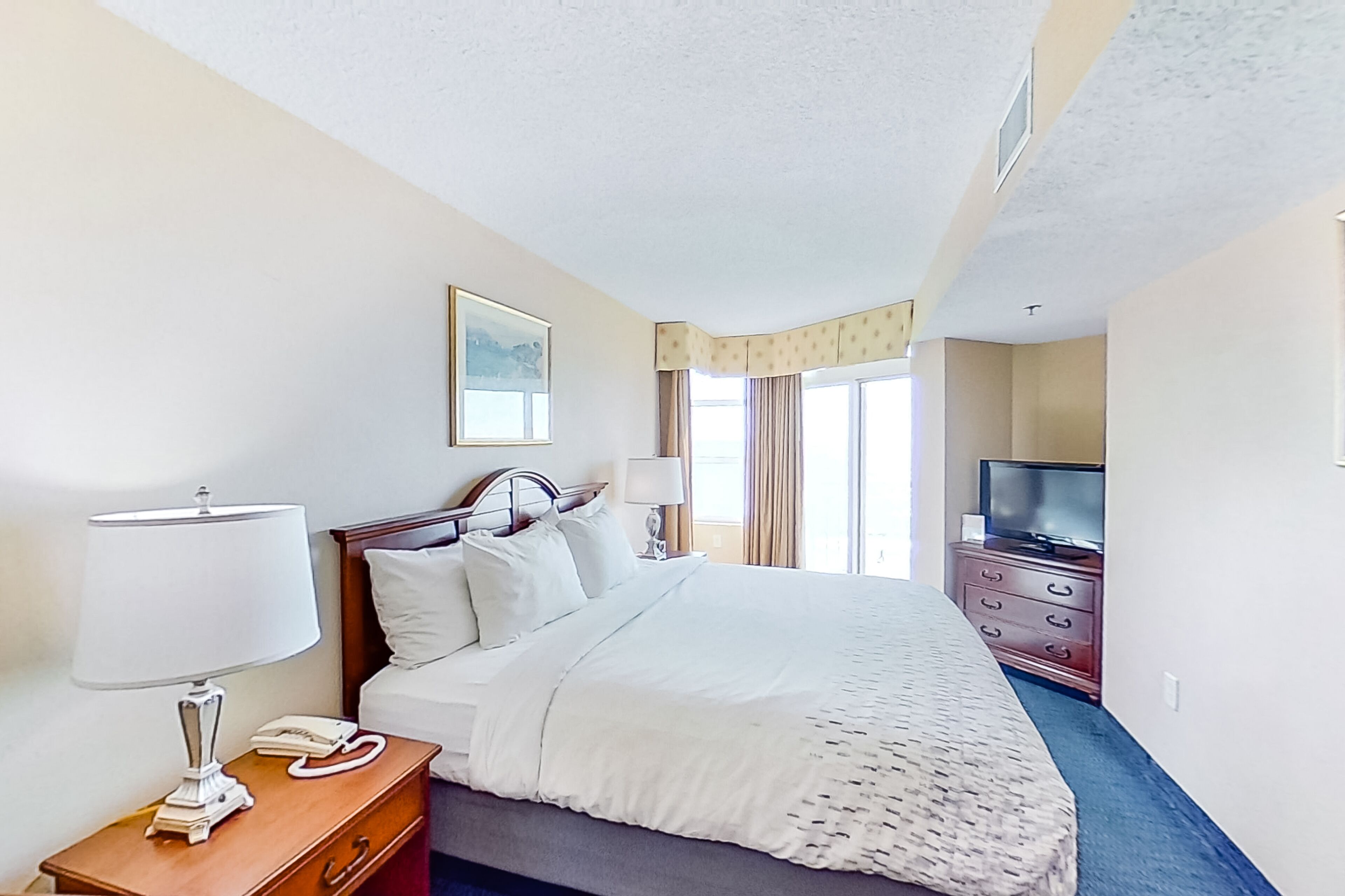 A hotel room with a bed, bedside table with a lamp, a telephone, a TV on a dresser, and a window with curtains.