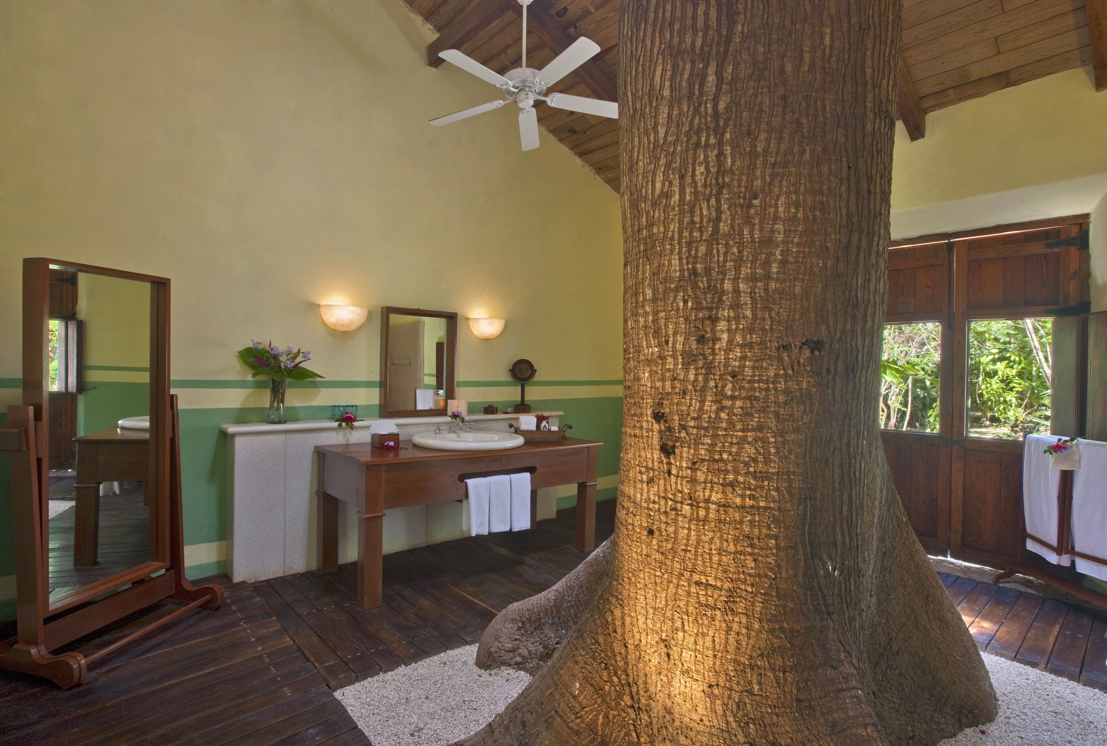 A bathroom with a large tree trunk centerpiece, wooden vanity, dual sinks, and a ceiling fan.