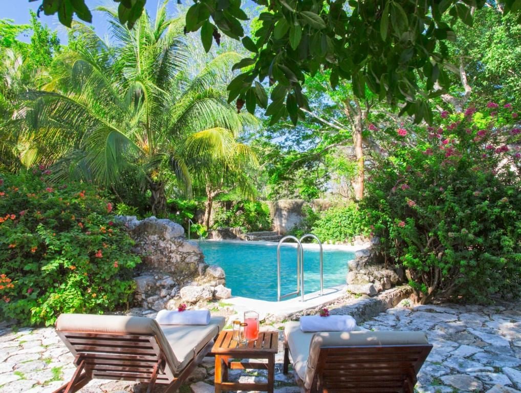 A poolside area with lounge chairs and a table, surrounded by lush greenery and a clear blue pool.