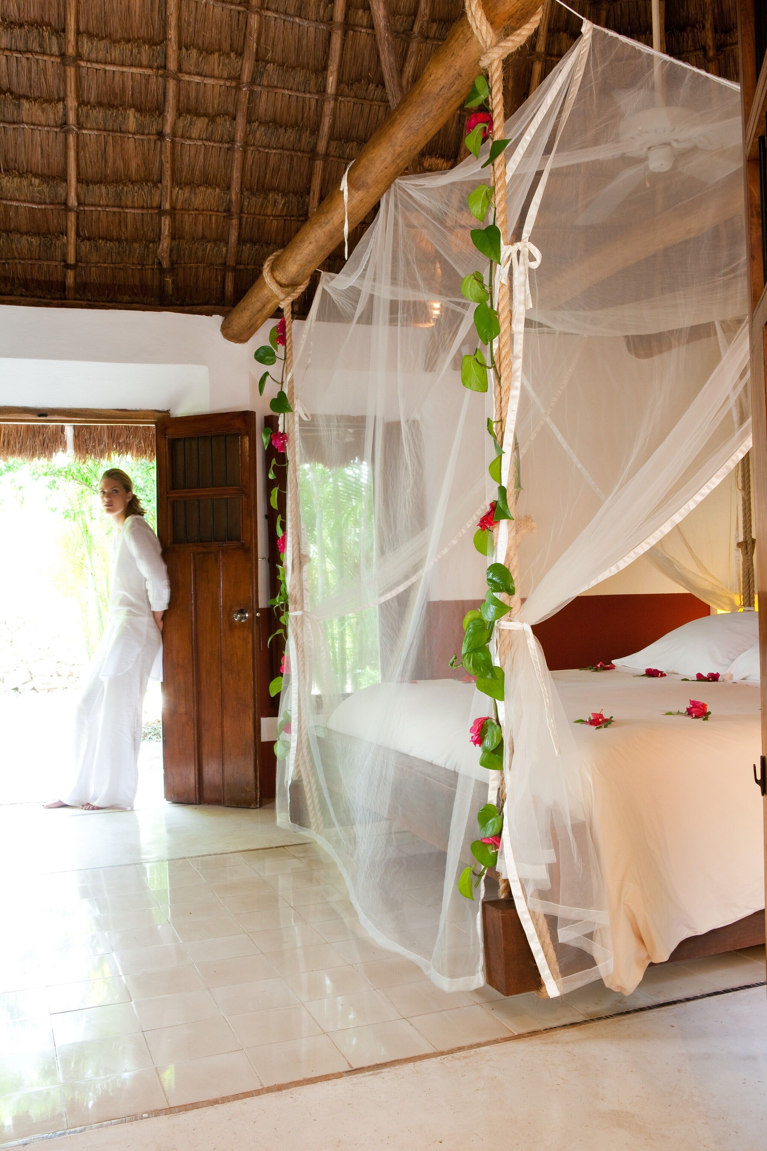 A room with a bed under a mosquito net, a wooden door, and a view of greenery outside.