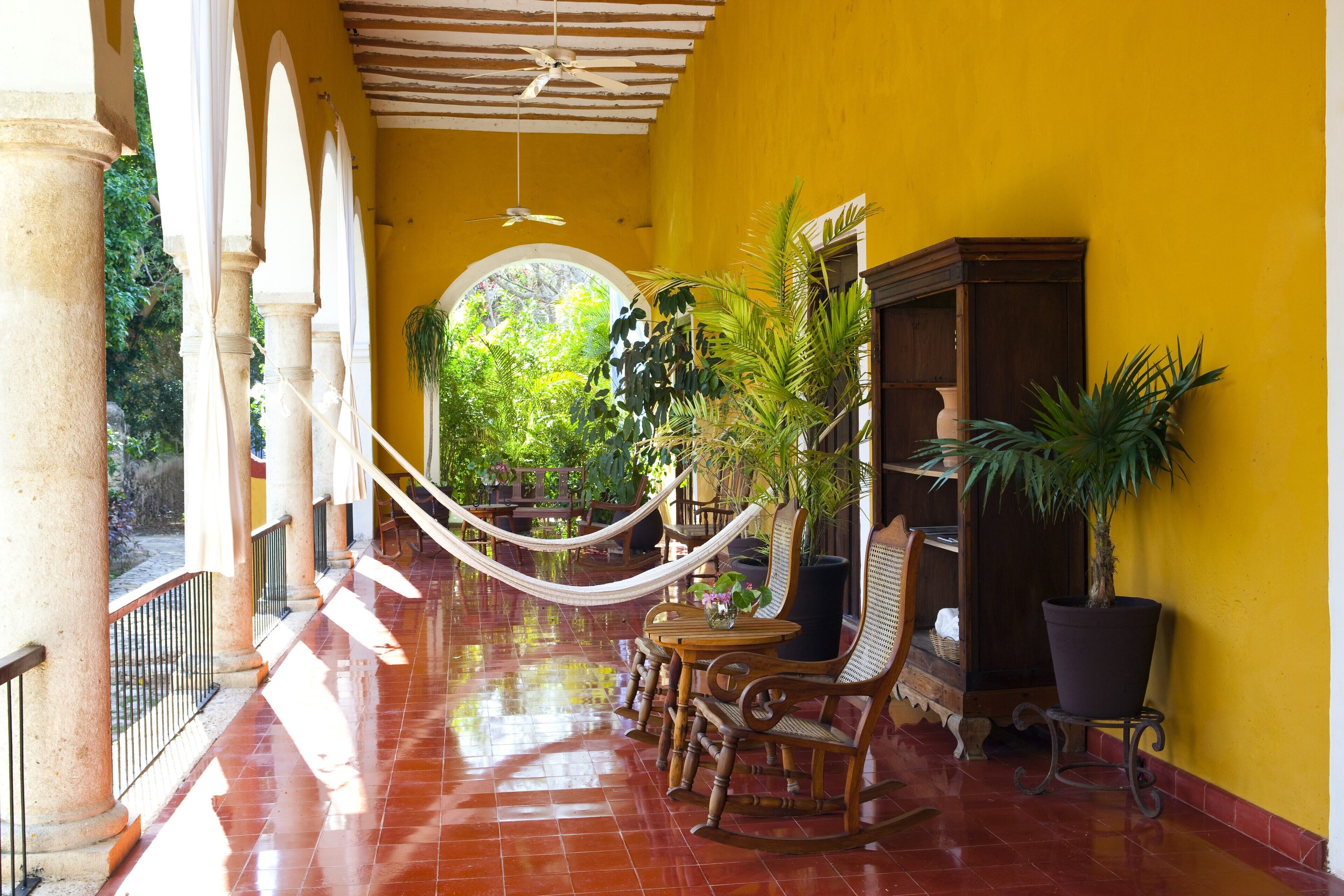 A yellow-walled corridor with a hammock, wooden furniture, and potted plants.