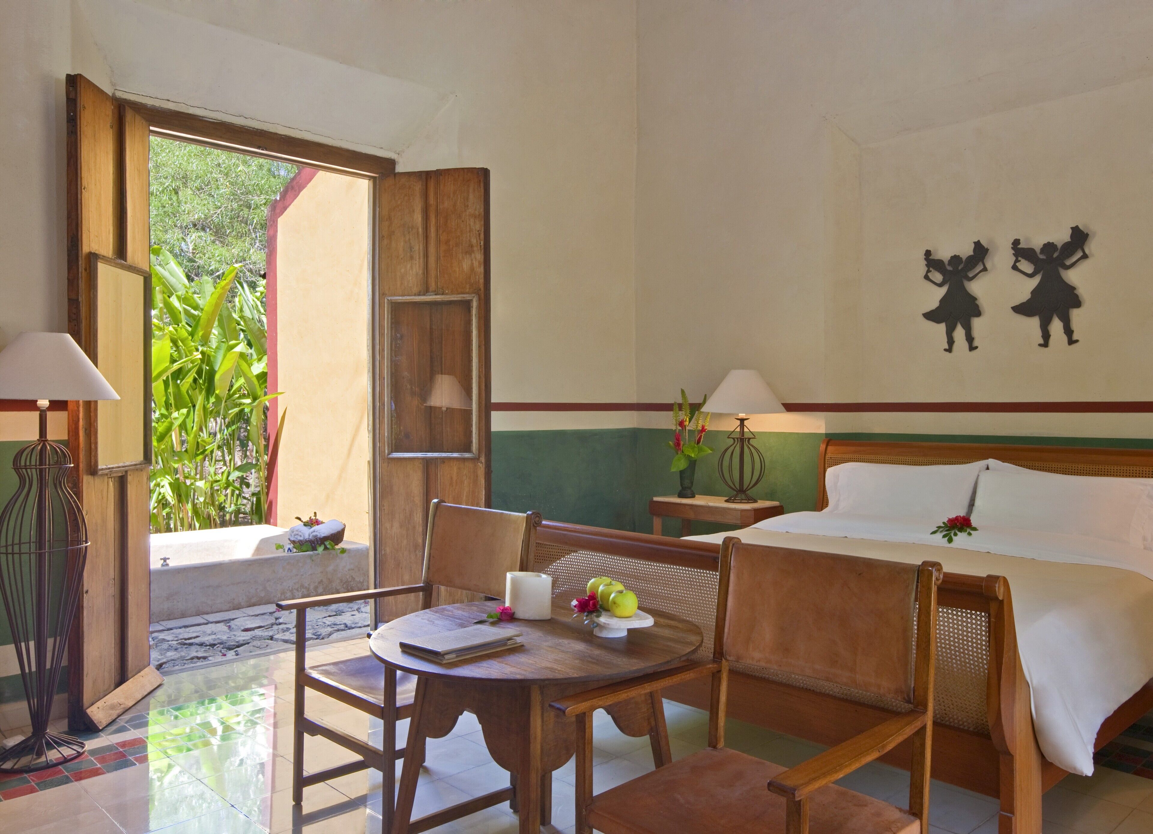 A hotel room with a bed, a wooden table with a notebook, a lamp, and a view of greenery outside.