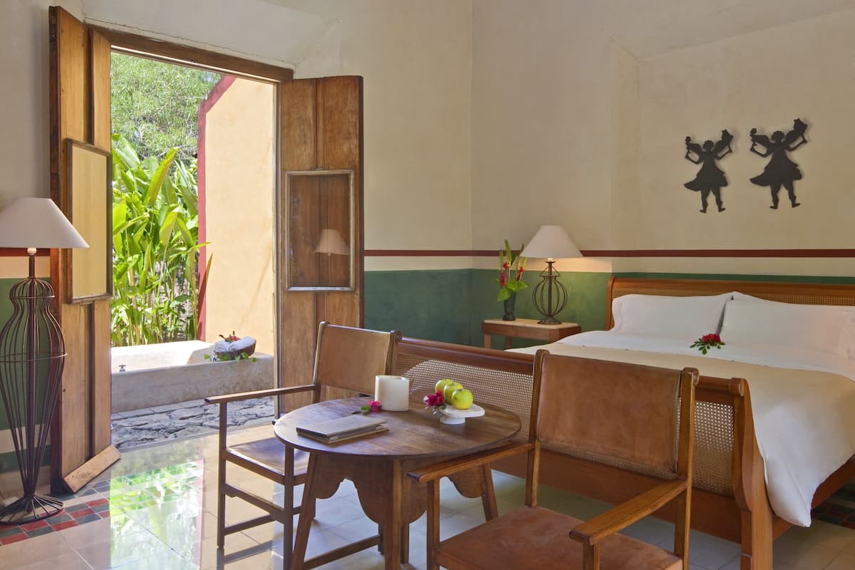 A hotel room with a bed, a wooden table with a notebook, a lamp, and a view of greenery outside.