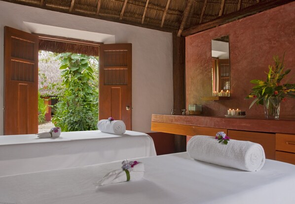 A hotel room with two beds, a wooden door, a mirror, a vanity with candles, and a view of greenery outside. - Hacienda San Jose Cholul (Cacalchén)