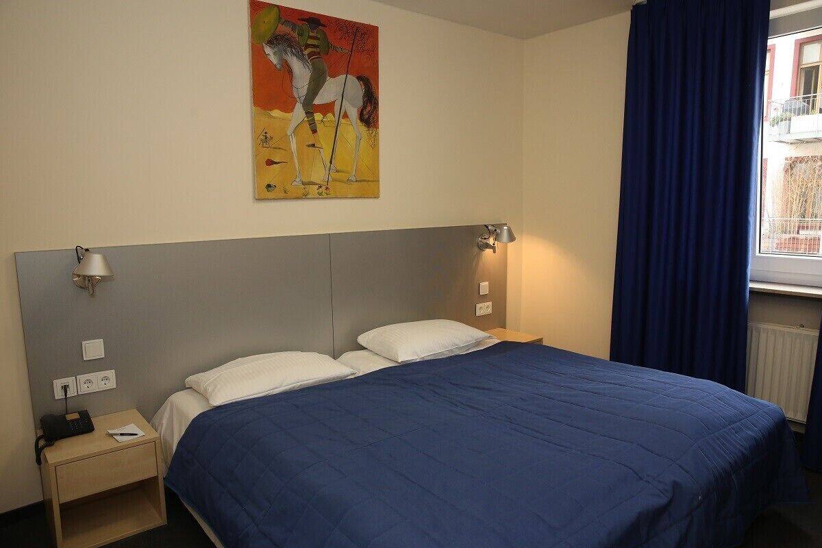 A hotel room with a bed, a nightstand, a lamp, a painting on the wall, and a window with curtains.