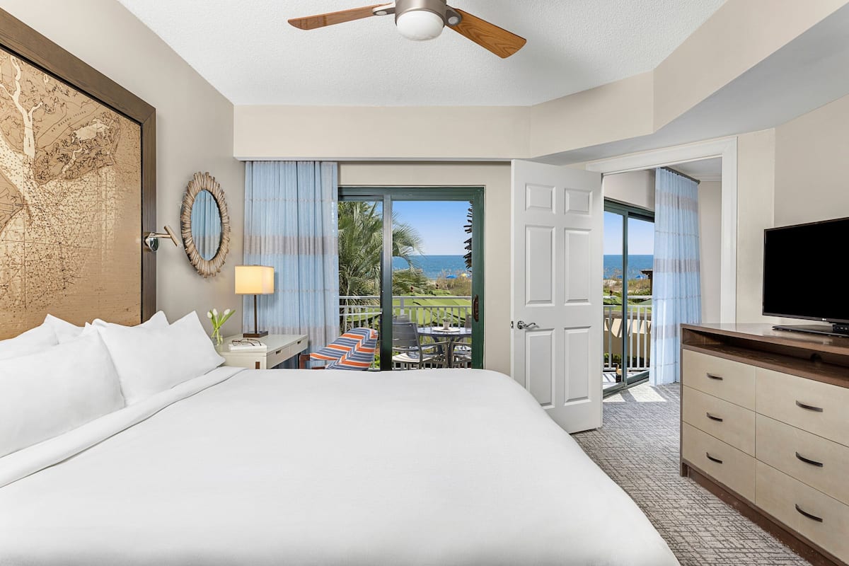 A hotel room with a large bed, a ceiling fan, a TV, a desk with a lamp, and a view of the ocean through a sliding door.
