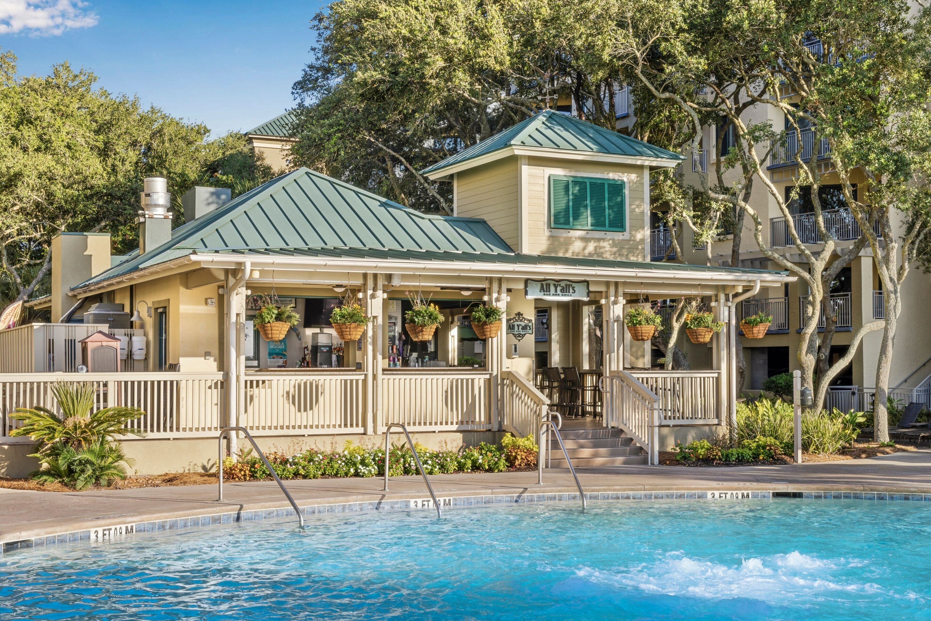 A poolside building with a green roof, a sign reading "All Y'all", and a pool with a tiled edge.