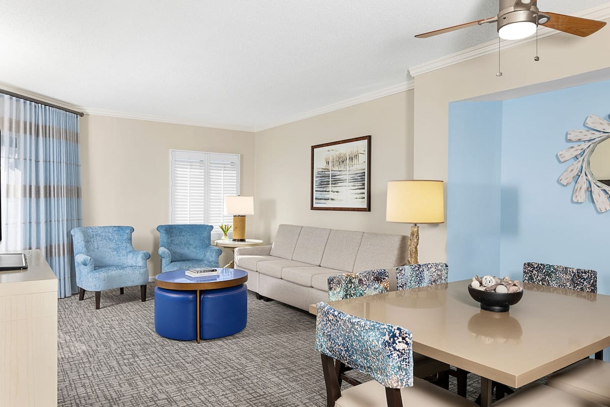 A hotel room with a sofa, two armchairs, a coffee table, a dining table with chairs, a ceiling fan, and a wall-mounted lamp.