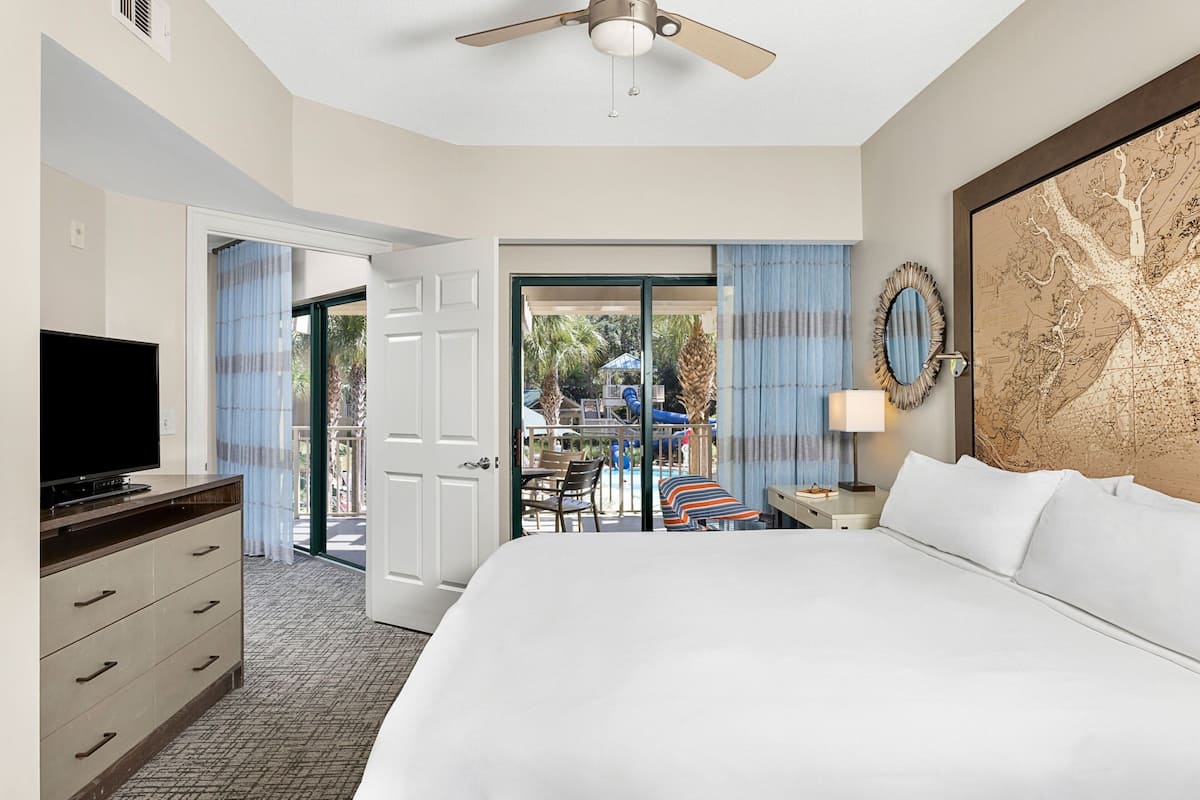 A hotel room with a large bed, a television, a desk with a lamp, and a view of an outdoor pool area.