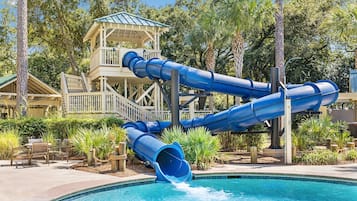 A pool area with a water slide structure, a covered seating area, and surrounding vegetation.