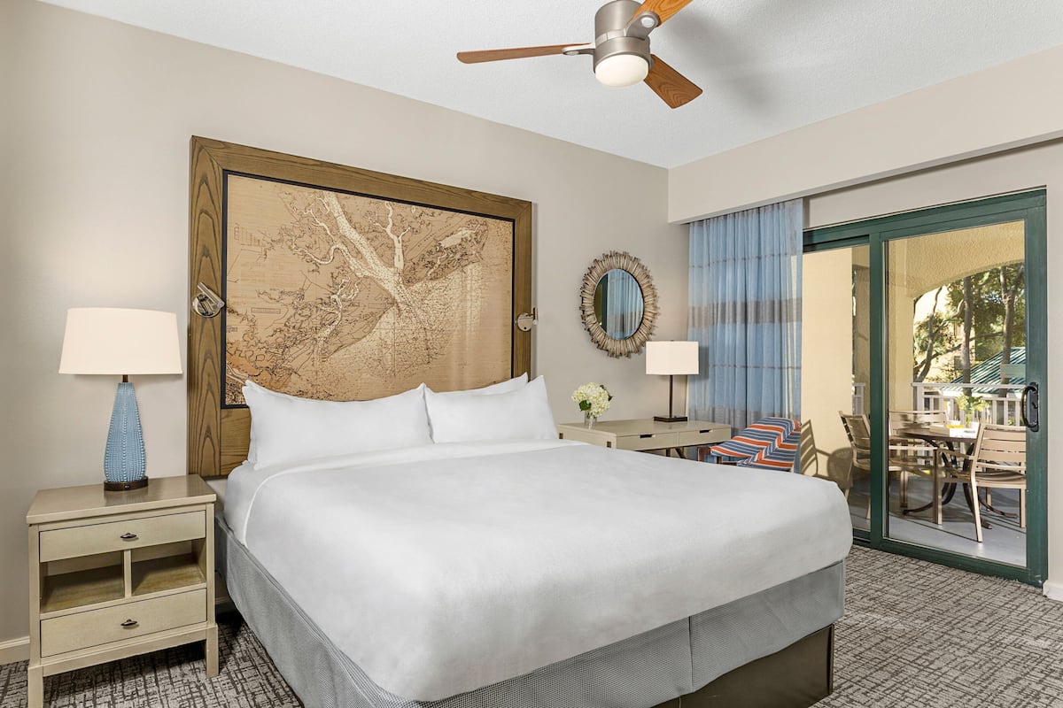 A hotel room with a large bed, a wooden headboard, two bedside tables with lamps, a mirror, and a view of an outdoor patio.