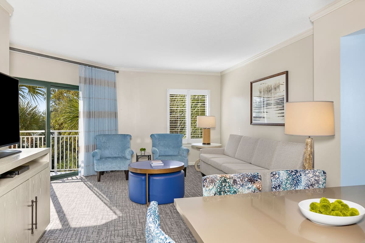 A modern hotel room with a balcony, a sofa, two armchairs, a coffee table, a TV, and a dining table with a bowl of fruit.