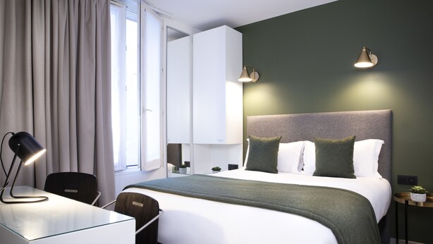 Een moderne hotelkamer met een groot bed, een bureau met een lamp, een stoel, een nachtkastje met een plant en een wandlamp.