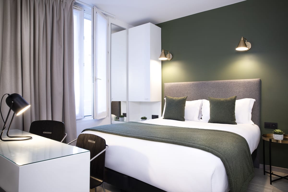Une chambre d’hôtel moderne avec un grand lit, un bureau avec une lampe, une chaise, une table de chevet avec une plante et une lampe fixée au mur.