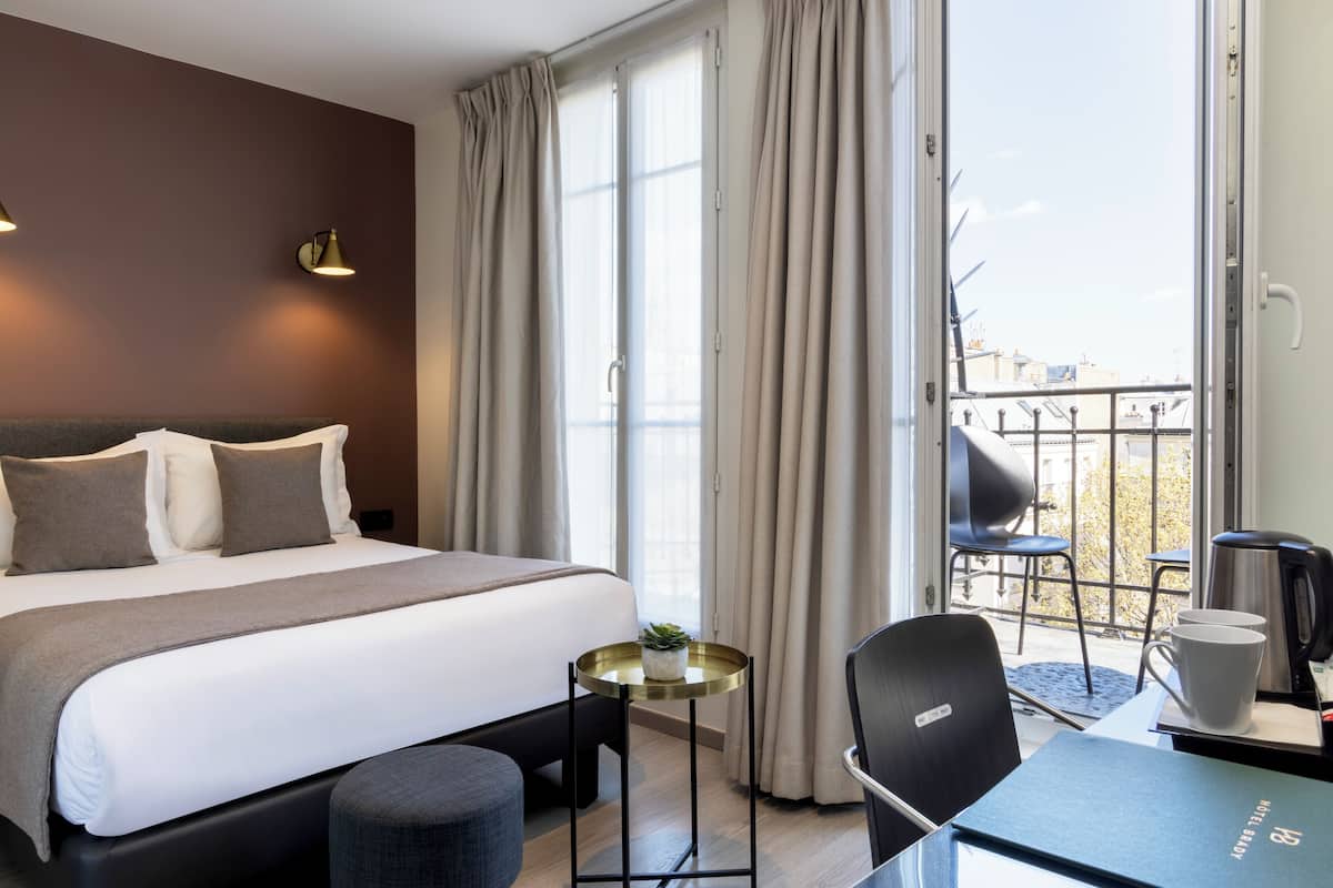 Une chambre d’hôtel avec un lit, une petite table avec une plante, une chaise, un bureau avec une machine à café et une vue sur le balcon extérieur.