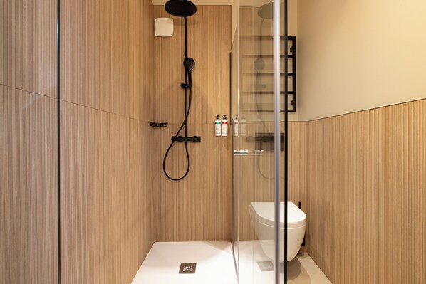A modern bathroom with a wooden panel wall, a glass shower enclosure, a white toilet, and a mounted shelf with toiletries.
