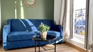 Een blauwe bank, een klein rond tafeltje met een plant, een ronde spiegel en een raam met gordijnen.
