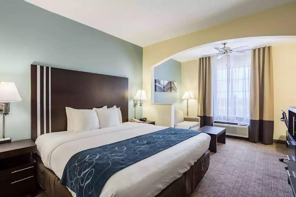 Une chambre d’hôtel avec un grand lit, deux tables de chevet, un bureau, une chaise, un ventilateur de plafond et une fenêtre avec des rideaux.