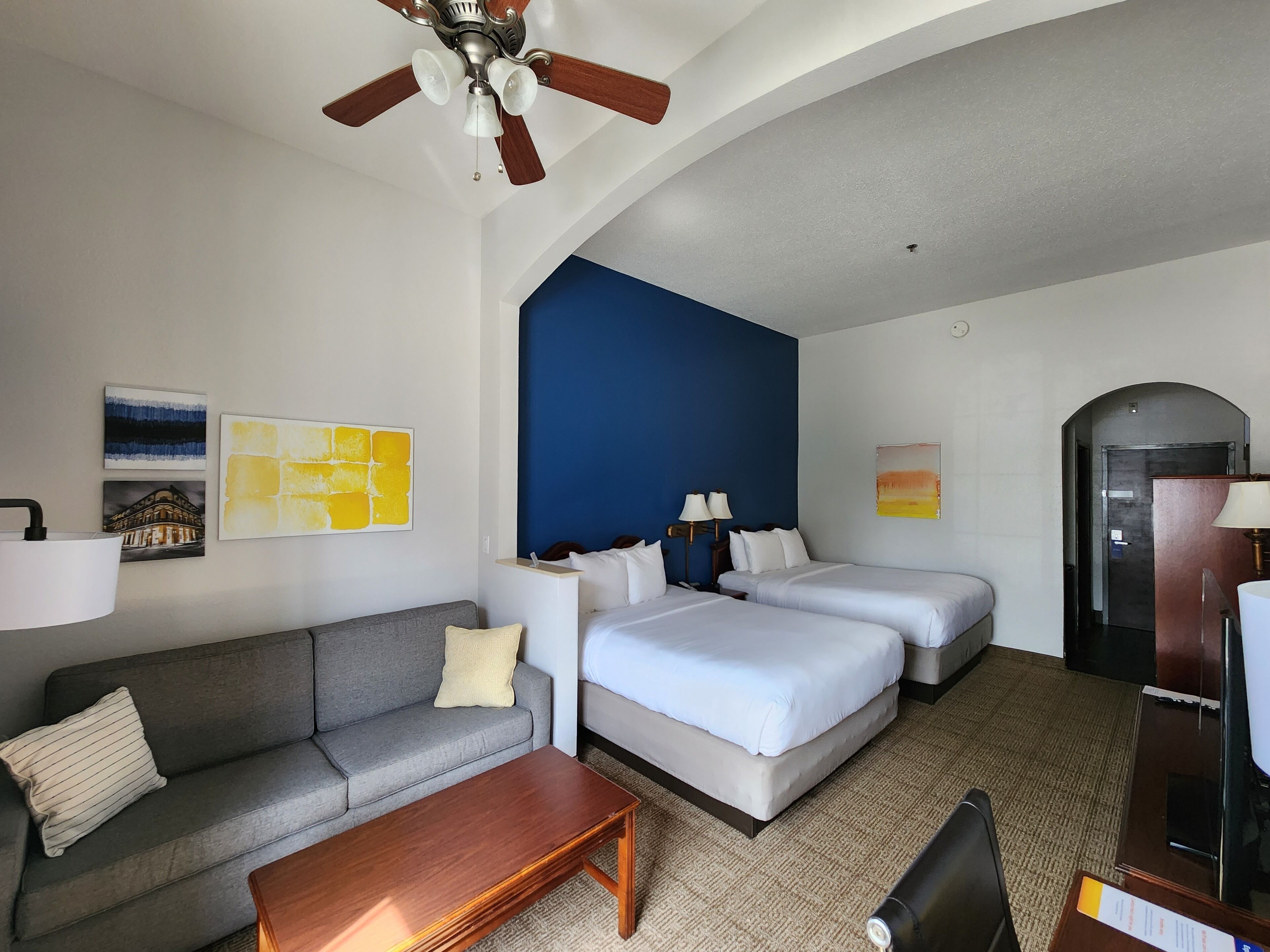 A hotel room with a gray sofa, two single beds, a wooden coffee table, a ceiling fan, and artwork on the walls.
