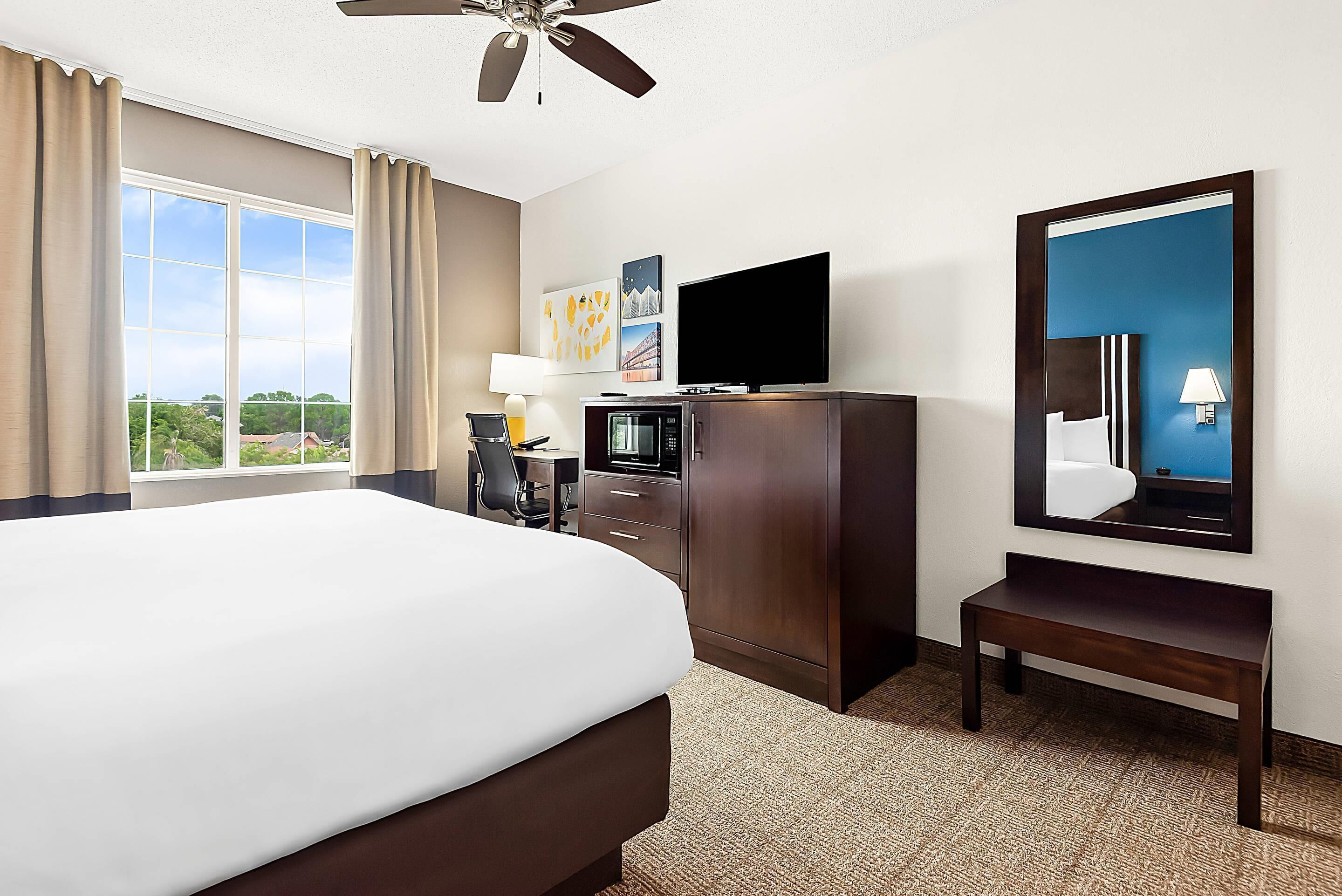 A hotel room with a large bed, a flat-screen TV, a desk with a chair, a mirror, and a view of the outdoors.