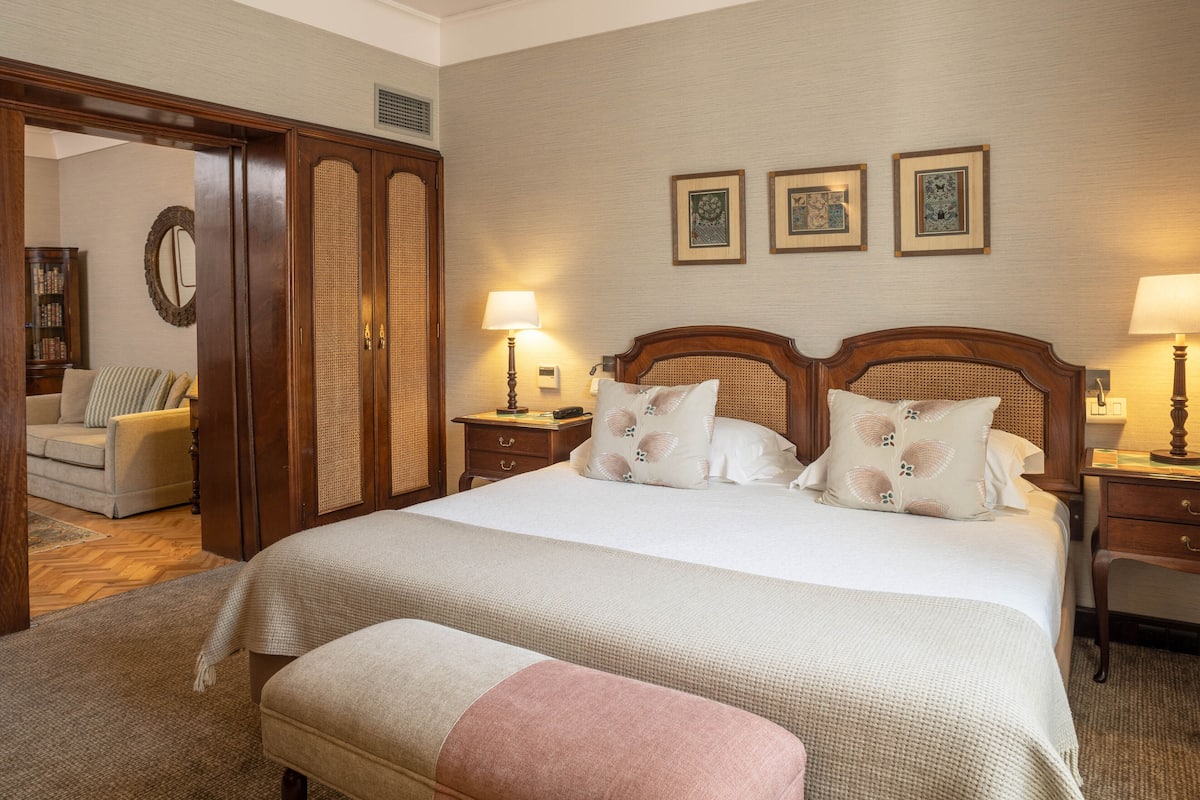 Una habitación de hotel con una cama grande, dos mesitas de noche, un armario de madera, un espejo y un sofá.