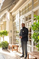 A man standing on a porch with a wooden bench, a potted plant, and a building with large windows and a beige facade.