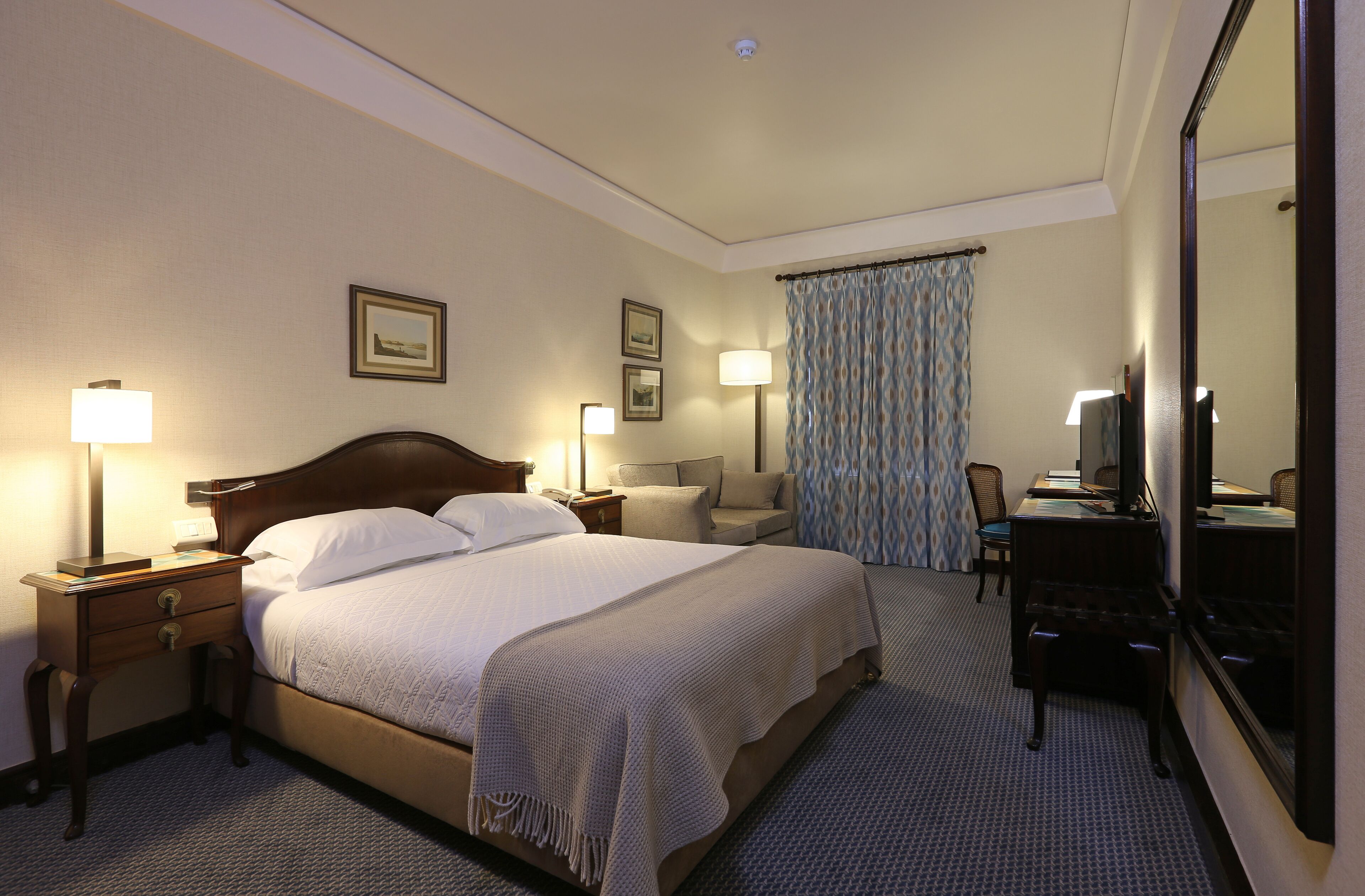 Ein Hotelzimmer mit einem großen Bett, Nachttischen mit Lampen, einem Schreibtisch mit Stuhl, einem Spiegel und einem gemusterten Vorhang.