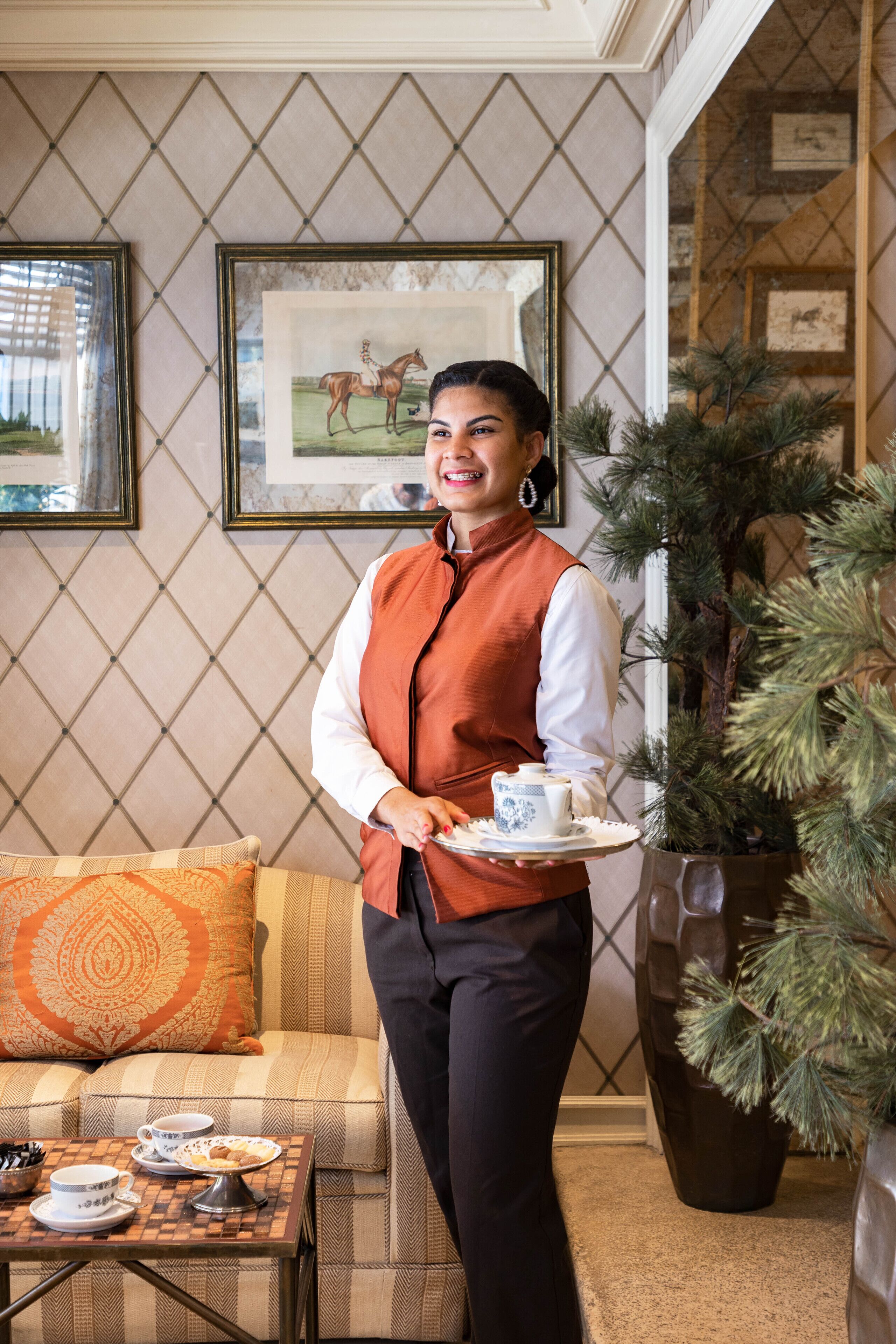 A person in a vest holding a tray with a teapot and cups, standing in a room with a patterned wall, a sofa, and a large potted plant.